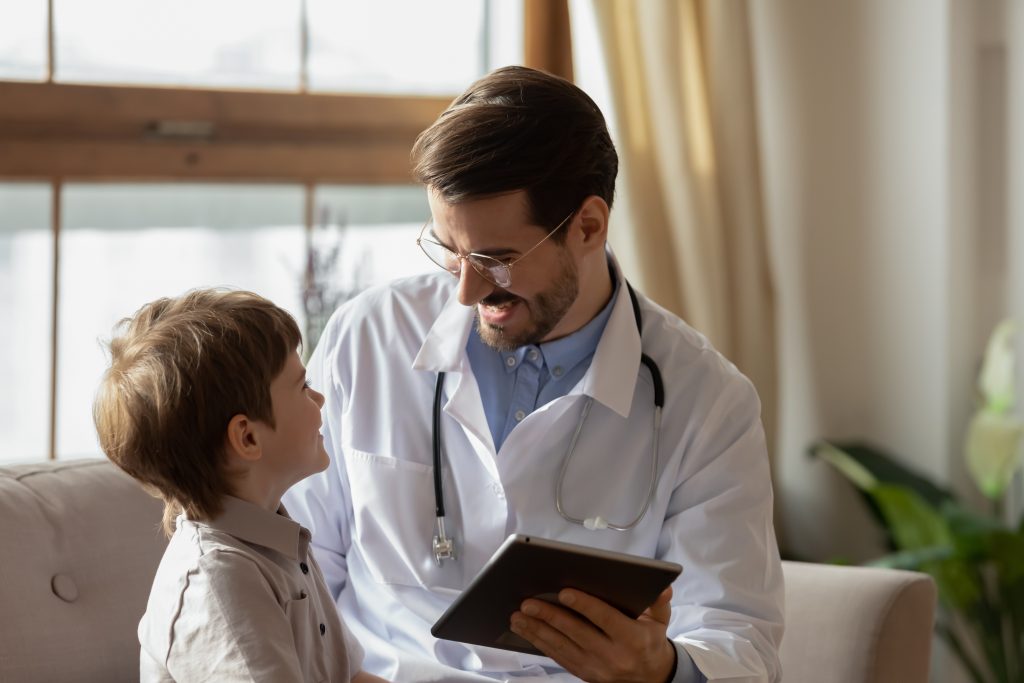 Smiling male pediatrician with little boy patient talk using tablet at consultation in hospital, caring doctor or GP do checkup speak with small child at clinic meeting, medicine, healthcare concept